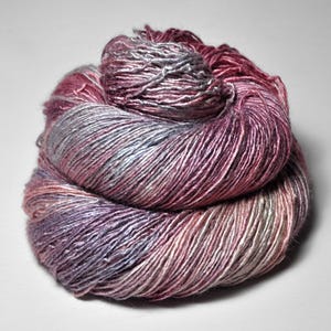 May include: A skein of yarn featuring a variegated color palette of deep magenta, soft lavender, and shimmering silver tones. The fiber has a distinct sheen and a twisted, multi-ply construction, wound into a compact, rounded bundle.