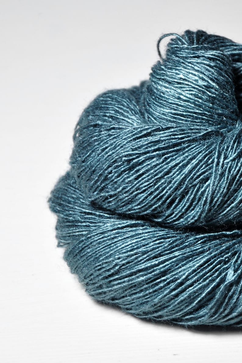 May include: A close-up of a ball of teal-colored yarn. The yarn has a textured, slightly shiny appearance, with individual strands visible. The yarn is tightly wound, creating a dense, rounded shape. Ideal for knitting and crochet projects.