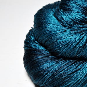 May include: A skein of deep teal yarn with a high-sheen, lustrous finish. The fibers are tightly twisted and densely packed, creating a rich, saturated color profile that reflects light across the textured surface of the individual strands.