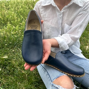 Op de afbeelding: Een paar marineblauwe leren instappers met een ronde neus en een gestikte zool. De schoenen worden getoond tegen een achtergrond van groen gras. Een persoon draagt een wit overhemd en een blauwe jeans.