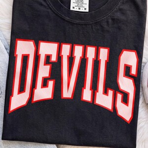 May include: A black short-sleeve t-shirt with a crew neckline featuring the word DEVILS printed in large, bold, white letters with thin vertical red stripes. The shirt has a white interior neck label with black text indicating size M.