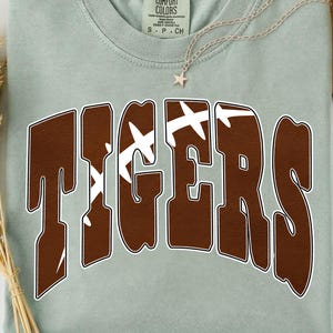 May include: A sage green short-sleeve t-shirt with a crew neckline and a Comfort Colors label. The front features the word TIGERS in large brown letters with a white outline, where the letters incorporate a white football lace graphic design.