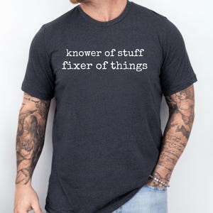 May include: Dark gray t-shirt with the text "knower of stuff fixer of things" in white. The shirt is a crew neck style and appears to be made of a soft material. The shirt is being worn by a person.