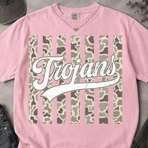 May include: A light pink short-sleeve t-shirt featuring a central graphic of vertical stripes with a brown and tan cow print pattern. Overlaid on the stripes is the word "Trojans" written in a white, distressed, cursive script font.