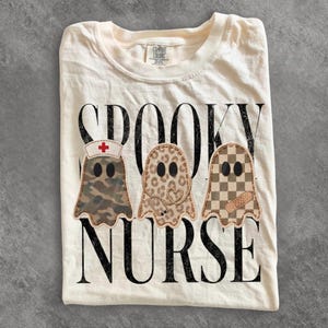 May include: An off-white t-shirt featuring the black text SPOOKY NURSE and three ghost illustrations. The first ghost has a camouflage pattern and a nurse cap, the second has a leopard print and stethoscope, and the third has a checkerboard pattern.