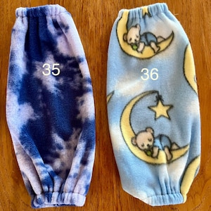 Pode incluir: Dois aquecedores de bra&ccedil;o de l&atilde; polar, um com um padr&atilde;o de tie-dye azul e branco e o outro com um padr&atilde;o azul e branco de ursos dormindo em luas. Os aquecedores de bra&ccedil;o s&atilde;o numerados 35 e 36.