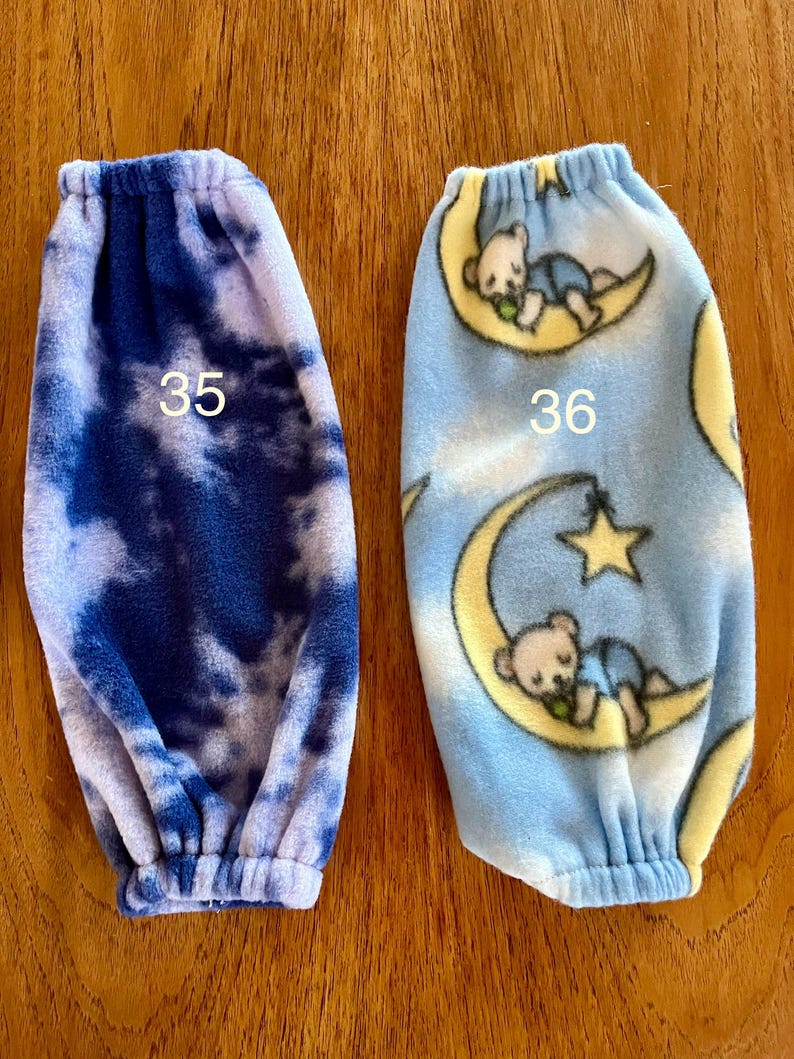 Pode incluir: Dois aquecedores de bra&ccedil;o de l&atilde; polar, um com um padr&atilde;o de tie-dye azul e branco e o outro com um padr&atilde;o azul e branco de ursos dormindo em luas. Os aquecedores de bra&ccedil;o s&atilde;o numerados 35 e 36.