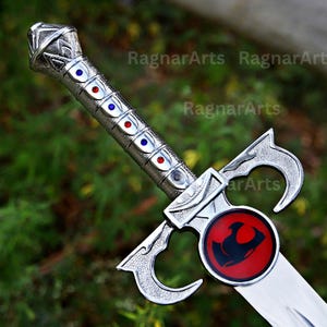 May include: A silver-toned sword featuring a textured pommel and a grip inlaid with alternating blue and red circular gems. The crossguard displays a prominent circular emblem with a black feline-like silhouette on a red background.