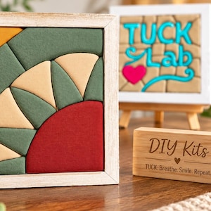 Puede incluir: Un marco de madera cuadrado que contiene un rompecabezas de tela geom&eacute;trico con segmentos en amarillo mostaza, verde oliva, crema y rojo oscuro. Al lado, un peque&ntilde;o bloque de madera grabado con el texto DIY Kits, TUCK. Breathe. Smile. Repeat.