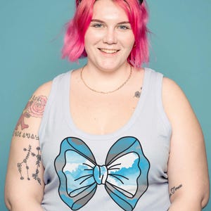 May include: A light blue tank top featuring a large bow design with a sky and cloud pattern. The bow is outlined in gray. The person is wearing a black and white bandana and has pink hair.