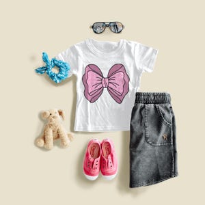 Könnte beinhalten: Ein weißes T-Shirt mit einer rosa Schleifen-Grafik, kombiniert mit einem grauen Jeansrock, rosa Turnschuhen, Sonnenbrille, einem blauen Scrunchie und einem Stoffhund. Die Turnschuhe tragen die Marke "Cliento".