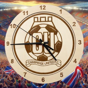 May include: A wooden clock featuring a brown and white Cambridge United F.C. logo. The clock face has black hands and white numbers.
