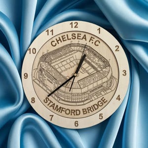 May include: A wooden clock featuring a black and white outline of Stamford Bridge, home to Chelsea Football Club. The clock face displays the numbers 1 to 12 and the text "Chelsea F.C." and "Stamford Bridge".