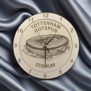May include: A white wooden clock with a black and white image of Tottenham Hotspur Stadium. The clock face has black numbers and the words "Tottenham Hotspur Stadium" are printed on the clock face.