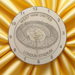 May include: A wooden clock with a white face and black numbers. The clock features a design of the London Stadium, home of West Ham United Football Club.