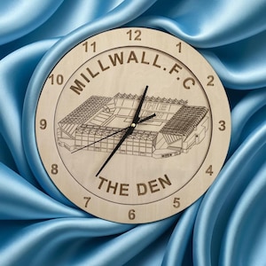 May include: A wooden clock with a light brown background featuring a black and white outline of a stadium with the text "MILLWALL.F.C THE DEN" around the perimeter. The clock face has black numbers and hands.