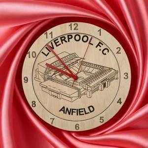 May include: A wooden clock featuring the text "LIVERPOOL F.C" and "ANFIELD". The clock face has a detailed illustration of a stadium. The clock hands are red, and the background is a red fabric.