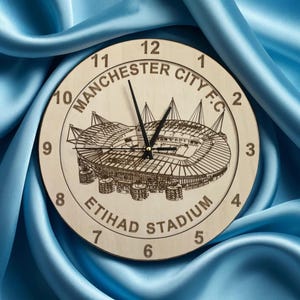 May include: A wooden clock with a black face and black hands. The clock face features a detailed image of the Etihad Stadium, home of Manchester City Football Club. The numbers 1 through 12 are printed around the edge of the clock face, along with the text "Manchester City F.C." and "Etihad Stadium".