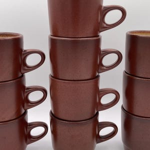 May include: A stack of twelve brown ceramic mugs with rounded handles. The mugs have a speckled texture and a slightly flared rim. The mugs are arranged in three stacks of four, creating a symmetrical display. The mugs are suitable for coffee or tea.