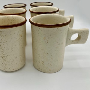 May include: Set of six speckled ceramic mugs. The mugs are off-white with brown rims and handles. The handles have a unique, rectangular design with a circular opening. The mugs are arranged in a semi-circle.