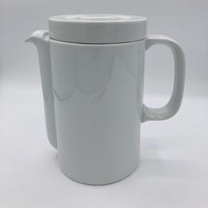 May include: A white ceramic teapot with a lid, spout, and handle. The teapot has a cylindrical body and a curved handle. The lid sits flush on top. The spout is curved and extends from the body.