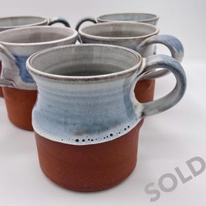 May include: Six handmade ceramic mugs. The mugs have a reddish-brown base and a light blue upper section with a white rim. Each mug has a curved handle. The word "SOLD" is visible in the lower right corner.