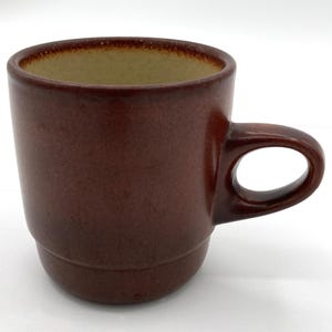 May include: A brown ceramic mug with a rounded handle. The mug has a dark brown exterior and a lighter brown interior. The mug is designed with a stacked base.