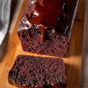 以下が含まれることがあります： 木製のまな板の上に、ダークチョコレートのフロスティングをかけたチョコレートケーキのスライスが、残りのパンの隣に置かれています。 ケーキは濃い茶色で、フロスティングは光沢があり滑らかで、側面には水滴があります。