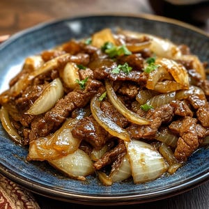 以下が含まれることがあります： 牛肉と玉ねぎの盛り合わせのクローズアップ。茶色の牛肉とキャラメル玉ねぎの柔らかい部分が特徴で、新鮮な緑のハーブで飾られています。食べ物は青いセラミックボウルで提供されます。
