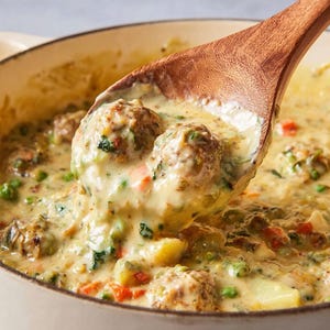 Peut inclure: Une cuillère en bois ramasse une soupe crémeuse avec des boulettes de viande, des petits pois, des carottes et des pommes de terre dans une casserole blanche. La soupe est jaune clair, et les boulettes de viande sont visibles avec des herbes vertes.