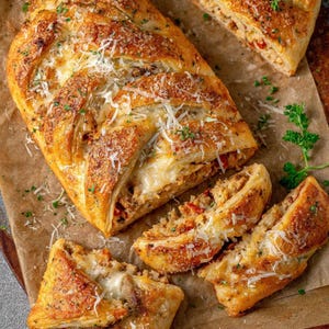 Puede incluir: Un pan trenzado dorado, cortado para revelar un relleno sabroso. El pan está cubierto con queso rallado y hierbas. Varias rebanadas se colocan alrededor del pan sobre una superficie marrón, mostrando la textura y los ingredientes del relleno.