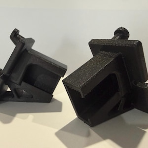 May include: Two black plastic brackets are displayed against a white background. The brackets have a rectangular shape with a C-shaped opening and a protruding screw. The brackets are designed to hold something in place.