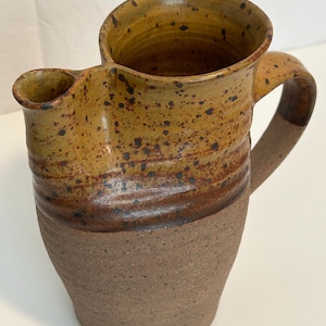 May include: A handmade ceramic pitcher with a unique spout and handle. The pitcher features a speckled, earthy color palette of brown, tan, and dark spots. The bottom half has a rougher texture, while the top is smoother.
