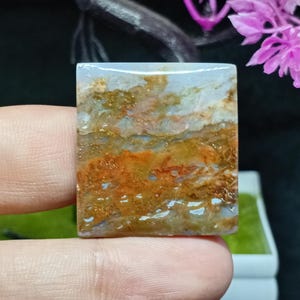 Puede incluir: Una gema rectangular con un patrón similar a un paisaje. La piedra presenta una mezcla de colores, incluyendo tonos de azul, naranja, marrón y blanco. La superficie pulida es lisa y reflectante, mostrando las variaciones naturales de color y textura.