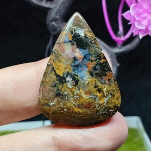 Puede incluir: Una gema pulida en forma de lágrima. La piedra presenta una mezcla de colores, incluyendo marrón, negro, amarillo y azul, creando un patrón único. La gema se sostiene en una mano, con un fondo borroso.
