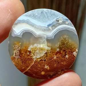 May include: A round, polished gemstone with a natural landscape pattern. The stone features layers of white, gray, and blue, with a base of brown and gold. The gemstone is held between two fingers.