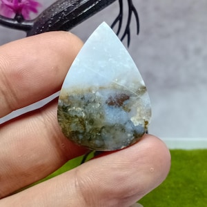 Puede incluir: Una gema en forma de lágrima con un degradado de color azul claro y marrón. La parte superior es azul pálido, que pasa a marrón y verde en la base. La piedra se sostiene entre los dedos, mostrando sus patrones y texturas naturales.