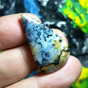 Pode incluir: Uma pedra preciosa de opala dendrítica em forma de lágrima com uma base translúcida azul-clara e branca, apresentando inclusões pretas complexas semelhantes a fetos e manchas de depósitos minerais amarelo-dourados na superfície polida.