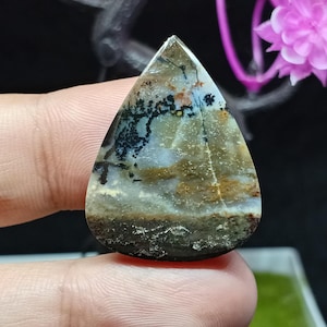 Puede incluir: Una gema en forma de lágrima con una superficie pulida. La piedra presenta un patrón similar a un paisaje con tonos marrones, negros y azules. El borde inferior tiene una apariencia oscura y texturizada. La gema se sostiene entre los dedos.