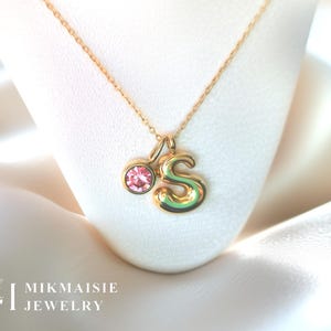 May include: A gold-coloured necklace with a pendant featuring the letter "S" and a pink gemstone. The pendant is suspended from a delicate chain. The necklace is displayed on a white surface.