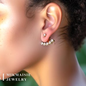 May include: A close-up of a person's ear adorned with two pieces of jewelry. The ear has a small, square stud earring and a gold-toned ear jacket with five clear, round stones. The jewelry is from the brand Mikmaisie Jewelry.