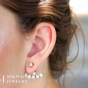 May include: A close-up of a silver earring set. The earring features a small, round stud and a decorative piece with five white, round stones. The earring is worn on the ear. The brand name "MIKMAISHI JEWELRY" is visible in the lower left corner.