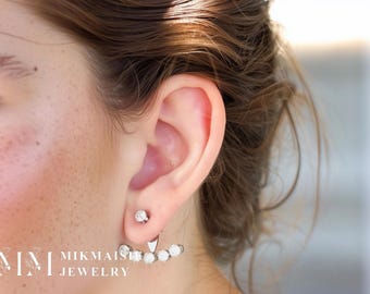 May include: A close-up of a silver earring set. The earring features a small, round stud and a decorative piece with five white, round stones. The earring is worn on the ear. The brand name "MIKMAISHI JEWELRY" is visible in the lower left corner.