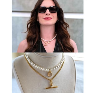 May include: A layered necklace set featuring a strand of white pearls and a gold-toned chain. The pearl strand includes a circular pendant, while the gold chain features a toggle bar closure. The set is displayed on a white neck form.