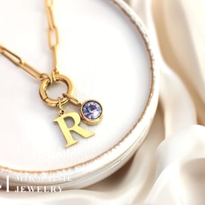 Op de afbeelding: Goudkleurige ketting met een grote schakelketting. De ketting heeft een bedel met de letter "R" en een ronde, lichtpaarse edelsteen. De sieraden worden gepresenteerd op een wit bord en een witte doek.