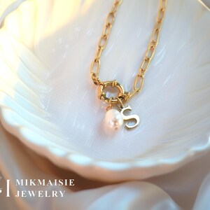 Peut inclure: Collier doré avec une chaîne à maillons larges et un fermoir circulaire. Une perle et la lettre "S" pendent du fermoir. Le collier repose sur un plat blanc en forme de coquillage. La marque "MIKMAISIE JEWELRY" est visible en bas à gauche.
