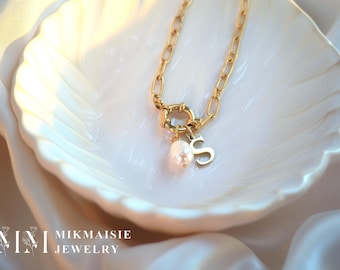 May include: Gold-coloured necklace featuring a chunky link chain and a circular clasp. A pearl and the letter "S" hang from the clasp. The necklace is displayed on a white, shell-shaped dish. The brand name "MIKMAISIE JEWELRY" is visible in the bottom left corner.