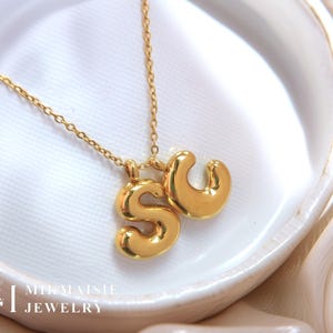 May include: A gold-coloured necklace with two initial pendants, featuring the letters "S" and "C". The pendants are in a bubble letter style. The necklace is displayed on a white surface.