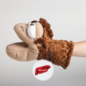 May include: Brown and tan plush hand puppet with a large eye and a stitched mouth. The puppet has a brown fur-like texture on the back of the hand. The puppet is designed to fit on a person's hand.  The puppet has a silicone lining inside the hand portion.