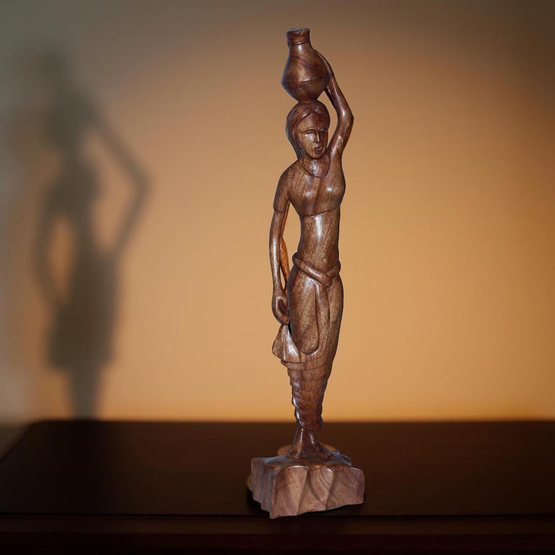 May include: A carved wooden sculpture of a woman carrying a water jug on her head. The figure is detailed with flowing garments and stands on a carved base. The wood has a rich, warm brown tone, and the sculpture is set against a neutral background.